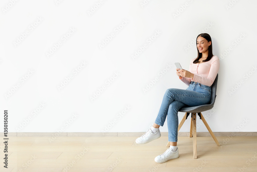 Mobile offer. Positive woman texting on cellphone, sitting on chair over white wall, enjoying free time and digital fun, full length Mobile offer. Positive woman texting on cellphone, sitting on chair over white wall, enjoying free time and digital fun, full length