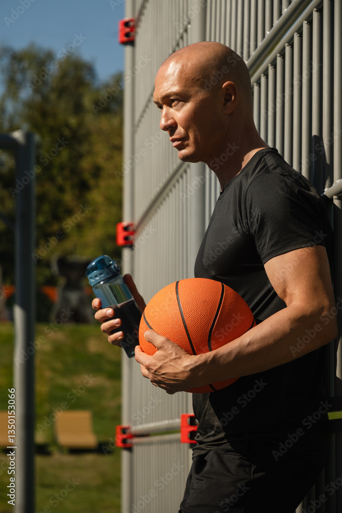 Portrait of basketball player or coach in black t-shirt holding basketball ball and water bottle on sports field. Outdoors. Basketball coach.