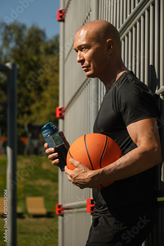 Obraz na płótnie Portrait of basketball player or coach in black t-shirt holding basketball ball and water bottle on sports field. Outdoors. Basketball coach. w salonie