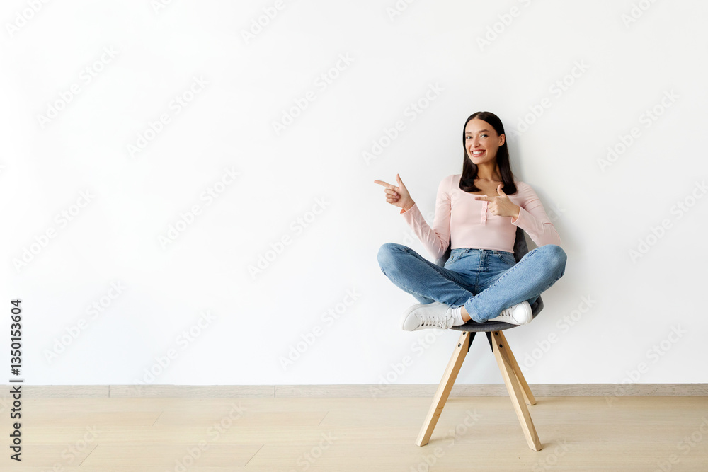 Cool promo. Happy European woman sitting in chair, pointing aside with both hands, offering empty space against white studio wall, mockup for your ad Cool promo. Happy European woman sitting in chair, pointing aside with both hands, offering empty space against white studio wall, mockup for your ad