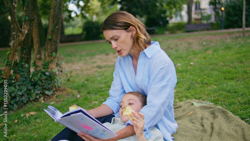 Fototapeta premium Picnic family reading book on green grass at weekend closeup. Kid eating apple
