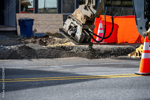 Roadworks covering trench dug for underground utilities with gravel and compacting with vibrating tamper attachment on excavator machine, new housing development construction project
