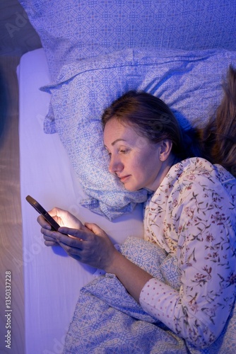  woman lies in bed in the moonlight, light from phone screen illuminating her face. Late night, tiredness inability to tear yourself away from gadget, atmosphere of loneliness dependence technology.