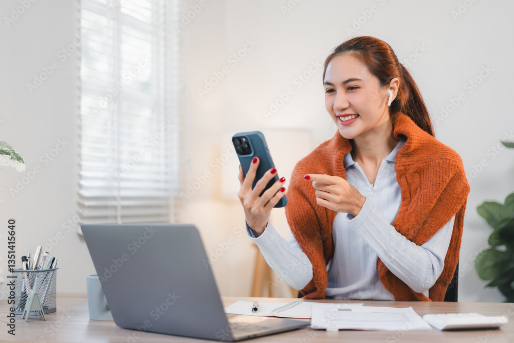 Asian businesswoman smiling while using smartphone and working on laptop in bright office setting. Asian businesswoman smiling while using smartphone and working on laptop in bright office setting.
