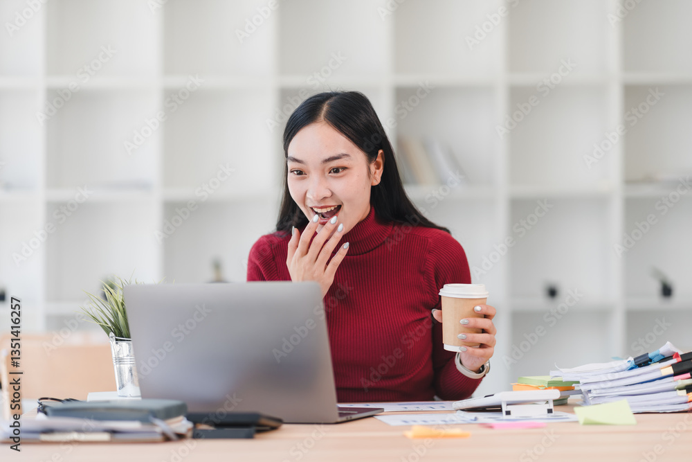 Asian businesswoman in red sweater smiling while working on laptop holding coffee cup in modern office setting. Asian businesswoman in red sweater smiling while working on laptop holding coffee cup in modern office setting.