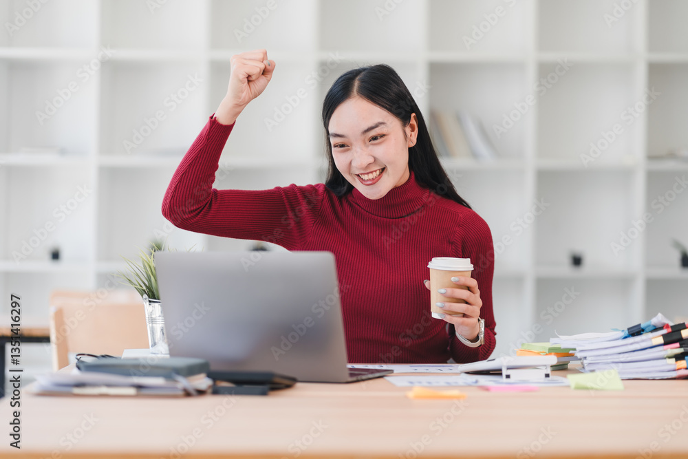 Obraz premium Asian businesswoman in red sweater celebrates success while holding coffee and working on laptop in modern office setting.
