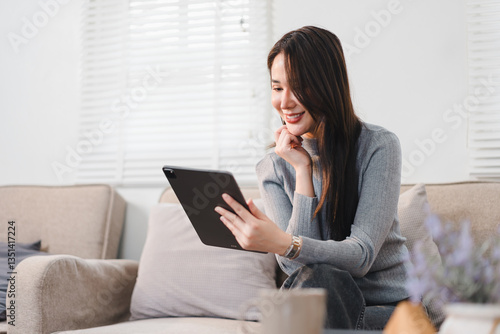 Cheerful Asian businesswoman sitting on sofa, using tablet in bright and cozy living room.