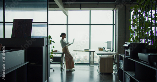 A young professional woman working on a laptop in a modern office with large windows and a scenic view, embodying focus, productivity, and a contemporary workspace atmosphere