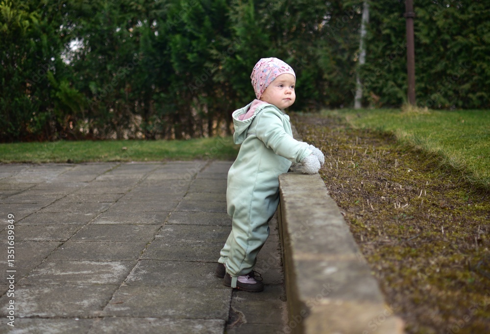 Obraz premium Baby girl in mint soft shell bodysuit is playing in a back yard in spring. Toddler girl walking on tiles footpath outside.
