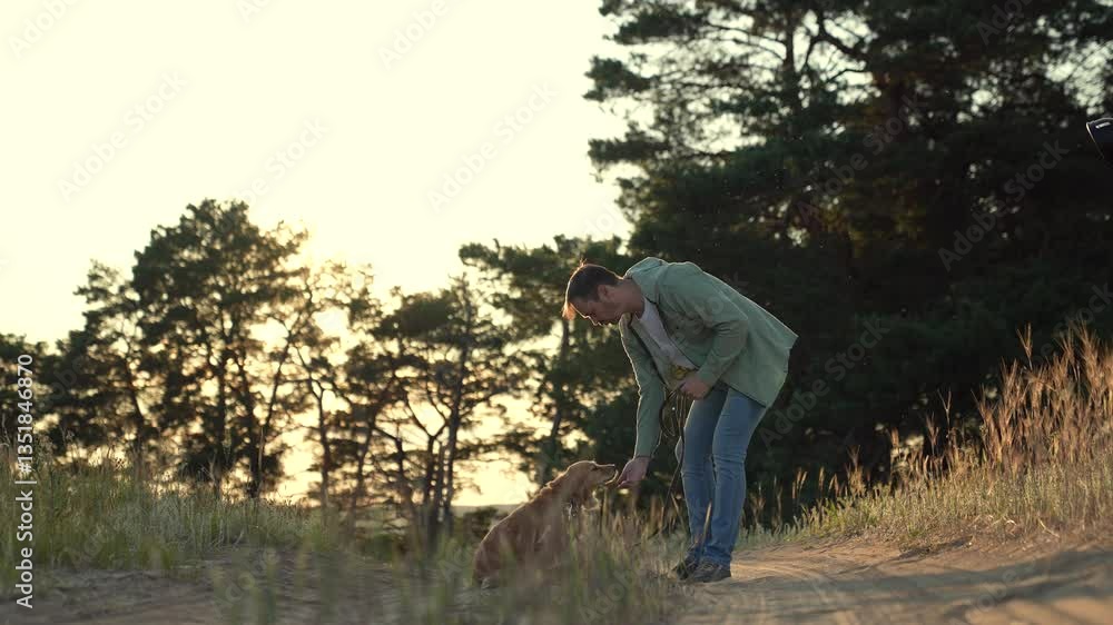Young man strokes red cocker spaniel dog on leash standing on park road man owner pets cute purebred spaniel dog walking at sunset in spring park man with dedicated spaniel dog in tranquil wild field Young man strokes red cocker spaniel dog on leash standing on park road man owner pets cute purebred spaniel dog walking at sunset in spring park man with dedicated spaniel dog in tranquil wild field