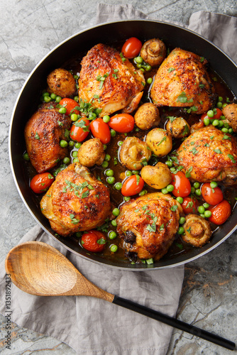 Homemade Chicken thighs baked with mushrooms, green peas, cherry tomatoes, garlic and herbs close-up in a frying pan on the table. Vertical top view from above
