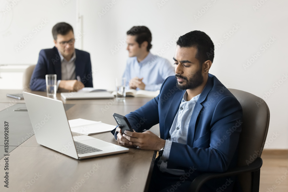 Focused Indian man working in office sit at table with laptop, holds smartphone, make business calls, check messages, conduct correspondence to client by emails, looks focused and productive. Workflow Focused Indian man working in office sit at table with laptop, holds smartphone, make business calls, check messages, conduct correspondence to client by emails, looks focused and productive. Workflow