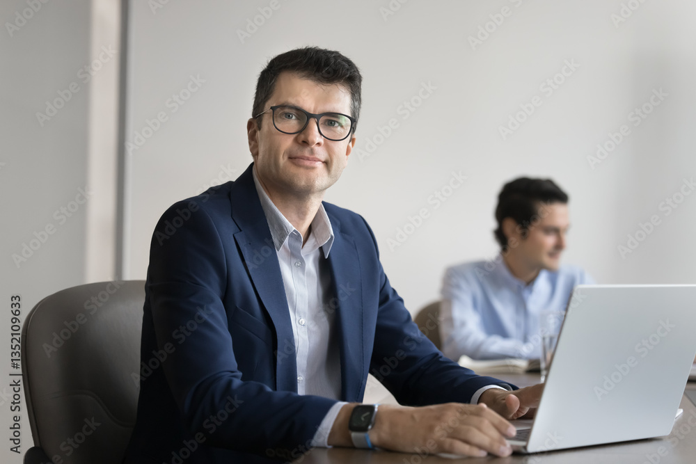 Successful professional middle-aged 45s businessman dressed in formal suit posing for camera with serious expression and confidence seated at desk with modern laptop. Corporate staff member portrait Successful professional middle-aged 45s businessman dressed in formal suit posing for camera with serious expression and confidence seated at desk with modern laptop. Corporate staff member portrait