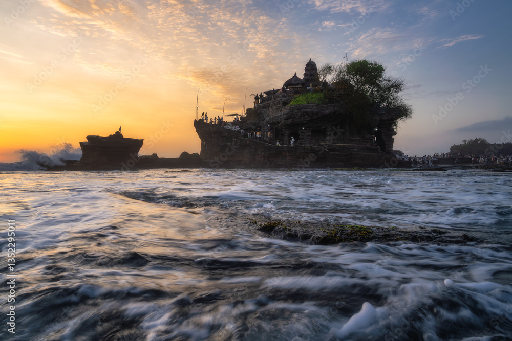 A stunning sunset fills the landscape with vibrant hues, casting a golden glow on a timeless Tanah Lot Temple, framed by soft ocean waves, creating a serene coastal scene, Bali, Indonesia