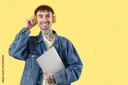 Young tattooed man in headphones with laptop listening to music on yellow bac...