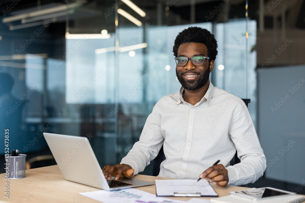 Obraz premium A happy, young professional works on a laptop, makes notes, and reviews data at his desk in a modern office with glass walls.