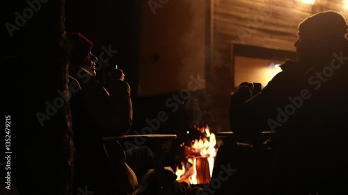 Couple sharing romantic kiss by campfire on chilly night,holding mugs and surrounded by warm firelight. Intimate moment is captured in cozy,winter outdoor setting.Perfect for love and holiday themes