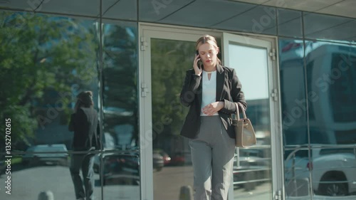 Lawyer exits mall on phone call with attentive expression, dressed in professional attire, holding handbag while walking, urban glass building reflects cityscape and vehicles