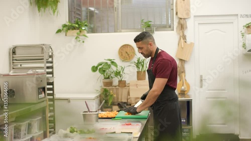 Professional chef slices pumpkin in a well-organized professional kitchen. The counter is set with meal prep containers for takeaway dishes. Warm, rustic ambiance with plants around. Wide angle, 4K