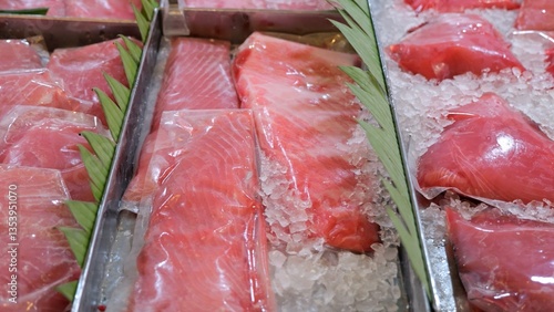 Tuna fillets and steaks, wrapped in plastic film, are displayed on beds of ice, garnished with green leaves, in metal trays at a fish market, emphasizing freshness and quality