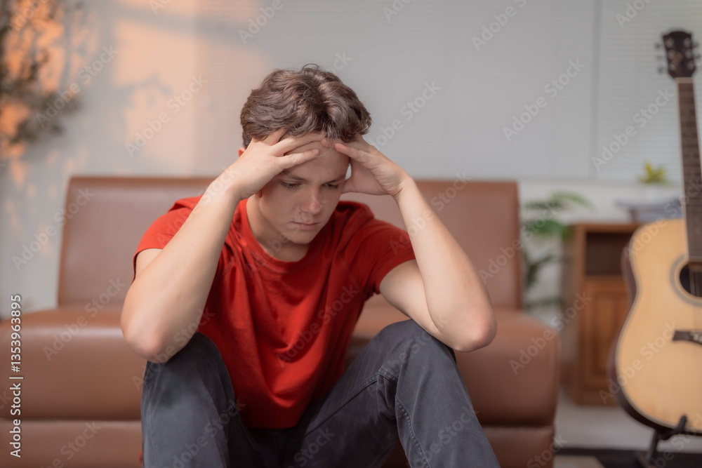 Young man holding head with hands, suffering from headache, sitting on floor at home, feeling exhausted, stressed and depressed, having migraine, emotional problem, teenage problems concept Young man holding head with hands, suffering from headache, sitting on floor at home, feeling exhausted, stressed and depressed, having migraine, emotional problem, teenage problems concept