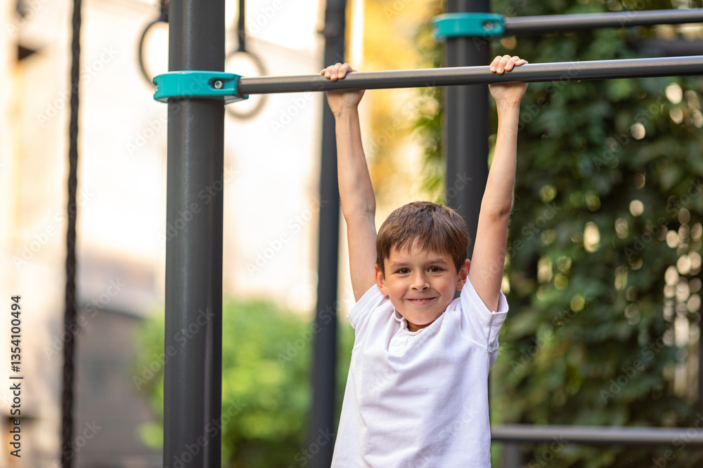 Little schoolboy, young athlete is pulling himself up on a sport horizontal bar on outdoor playground on sunny day. Active childhood, acrobat, workout on backyard, summer camp concept Little schoolboy, young athlete is pulling himself up on a sport horizontal bar on outdoor playground on sunny day. Active childhood, acrobat, workout on backyard, summer camp concept