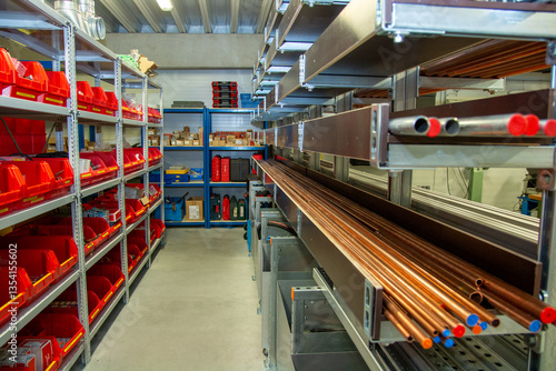 Industrial storage room with organized metal pipe racks and plastic bins filled with various components, tools, and parts for plumbing, HVAC, or mechanical system assembly.
