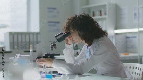 Young female scientist wearing lab coat and badge, analyzing samples under microscope and taking notes at desk with test tubes and flasks on it, doing laboratory research