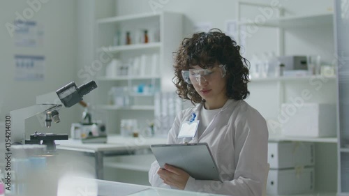 Wallpaper Mural Young female scientist in white coat and safety glasses sitting at desk with microscope, using digital tablet during workday in modern medical laboratory Torontodigital.ca