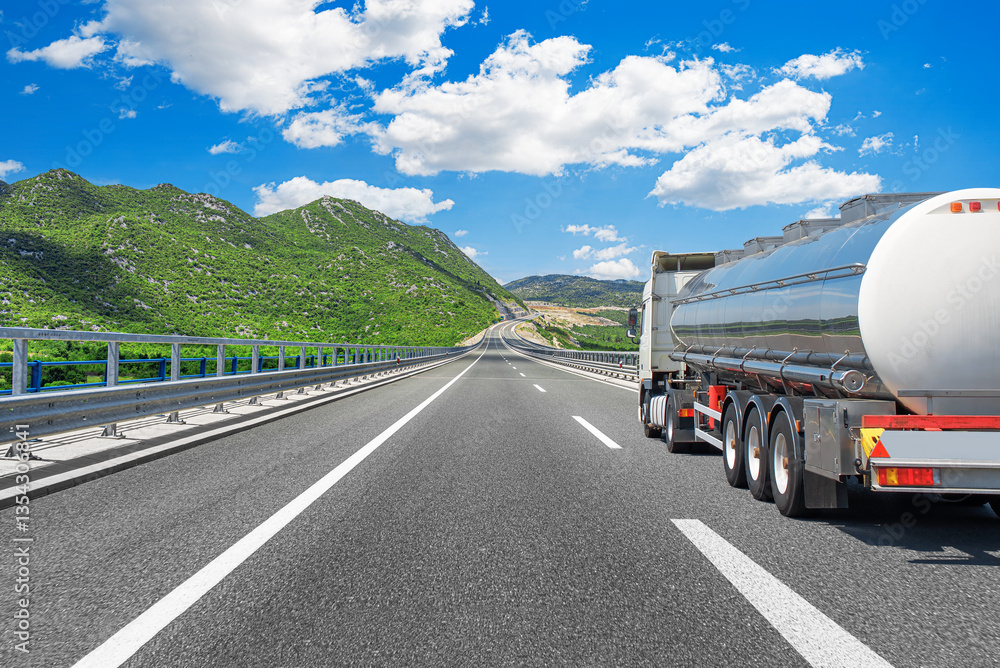 A shiny fuel tanker truck travels along a well-maintained highway surrounded by green mountains under a bright blue sky with scattered white clouds.