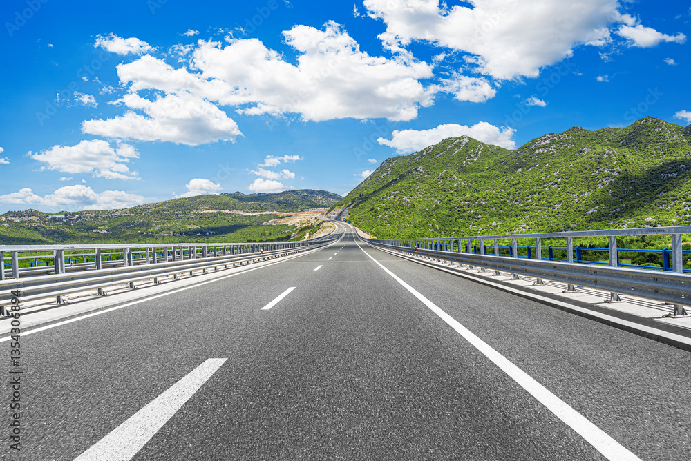 A modern, well-maintained highway stretches into the distance, surrounded by lush green mountains under a bright blue sky with scattered white clouds.