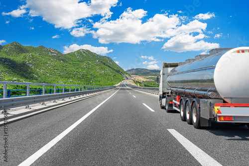 Obraz na płótnie A shiny fuel tanker truck travels along a well-maintained highway surrounded by green mountains under a bright blue sky with scattered white clouds. w salonie