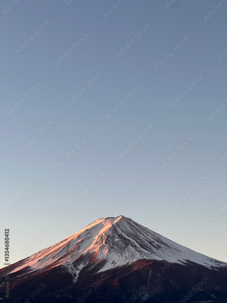 Fototapeta premium A stunning view of Mount Fuji at sunrise with golden sunlight illuminating the snow-capped peak, set against a clear blue sky in Yamanashi Prefecture, Japan.