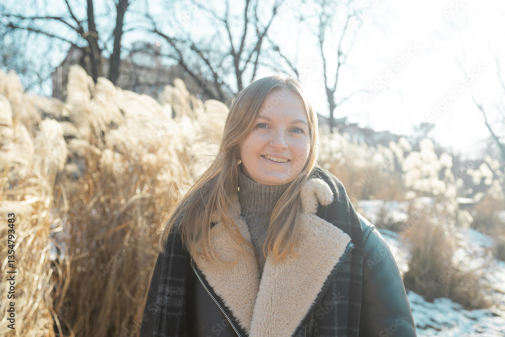 Young blonde cheerful girl in stylish jacket, looking at the camera smiling on a winter day. Close-up delicate white pampas grass swaying in wind. High quality photo