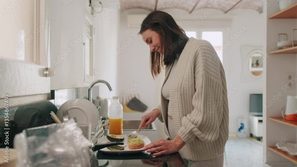 A woman smiles while preparing and eating avocado toast for breakfast in her kitchen. She's wearing casual comfy clothing. A woman smiles while preparing and eating avocado toast for breakfast in her kitchen. She's wearing casual comfy clothing.