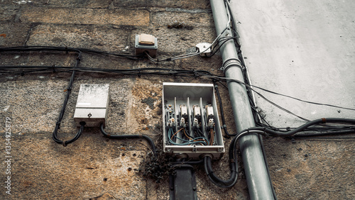 Close-up of an old electrical distribution box with open cover and exposed wiring mounted on a weathered stone wall, along with cables and conduit pipes, outdoors during daytime
