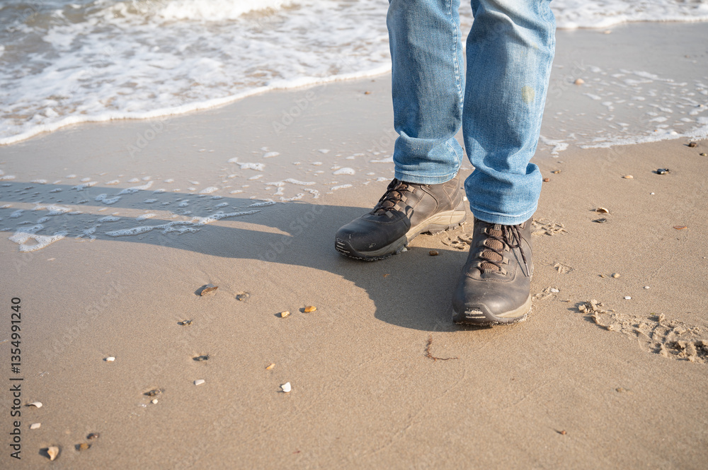 legs of a man in blue jeans, standing on a beach in hiking boots