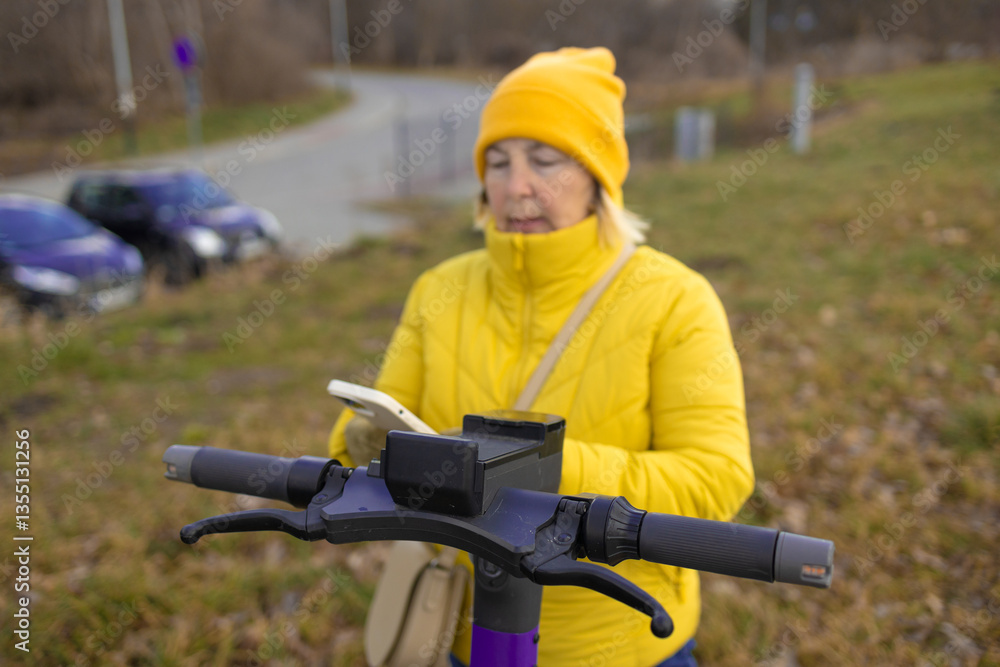 A woman in a yellow jacket and beanie rents an electric scooter using her phone in an urban park. The scene captures the convenience of app-based rental services for eco-friendly transportation.