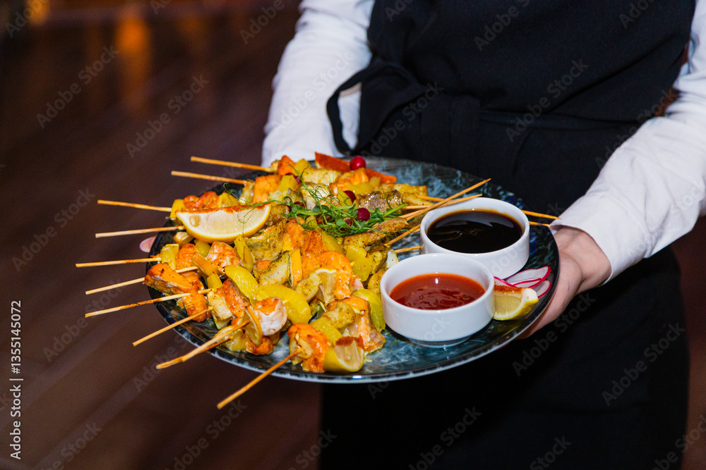 Shish kebab from seafood and red fish on wooden skewers on a plate with sauces in the hands of the waiter. overall plan