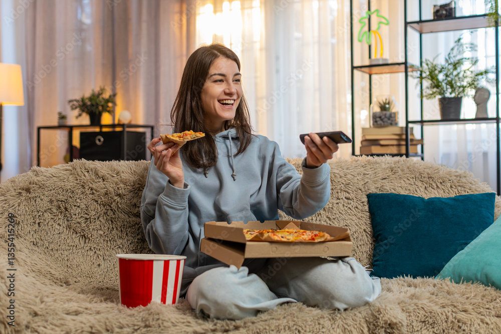 Obraz premium Happy young Caucasian woman eating delicious junk food pizza and popcorn sitting on sofa couch watching TV show or movie in home living room. Adult girl in casual clothes enjoying weekend in apartment