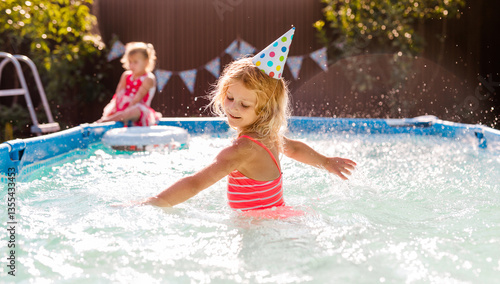 Curly-haired blonde girl splashing water in the pool during a party, wearing a paper party hat