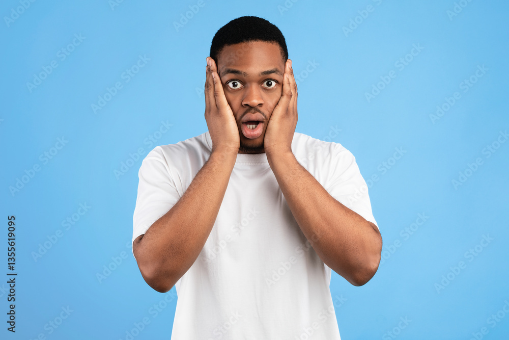 Fototapeta premium Overwhelmed Black Man Cupping Face In Hands Looking At Camera In Shock Standing On Blue Background. Studio Shot