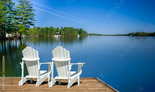 Two white Adirondack chairs on a wooden dock face the calm Muskoka lake, its glassy surface from a long exposure reflecting the sky. Cottages among green trees add to the serene scene.