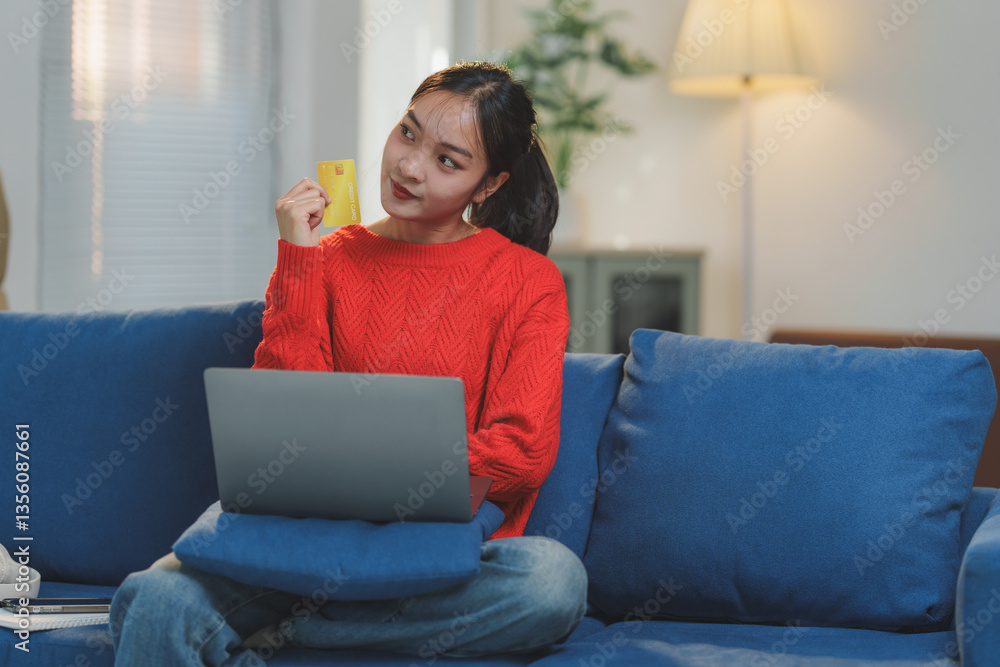 Young woman sitting on a blue sofa, holding a credit card while using a laptop. She appears thoughtful, possibly considering an online purchase in a cozy living room setting Young woman sitting on a blue sofa, holding a credit card while using a laptop. She appears thoughtful, possibly considering an online purchase in a cozy living room setting