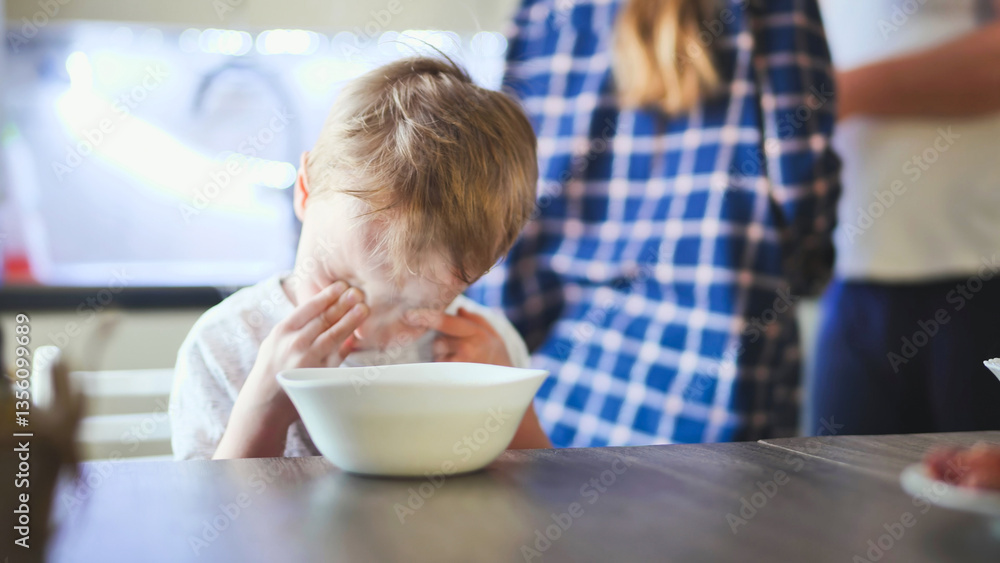 Sleepy toddler rubbing eyes while eating, parents watching nearby in sunlit kitchen interior Sleepy toddler rubbing eyes while eating, parents watching nearby in sunlit kitchen interior