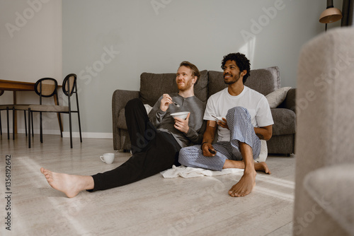 Принти на полотні In a living room, a White brown-haired man and a Black brunet man, a gay couple in their 20s wearing home clothes, are sitting on the floor resting their backs on a sofa, eating, and watching TV