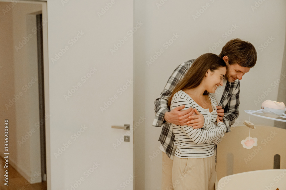 A White father in his late 30s, hugging his mid-30s White wife from behind over her shoulders as they both look into a cute crib while standing inside of a white room, with a door beside them A White father in his late 30s, hugging his mid-30s White wife from behind over her shoulders as they both look into a cute crib while standing inside of a white room, with a door beside them