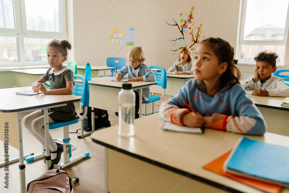 A young, school-aged White boy who is dressed in a button-up shirt, sitting at his desk in a first-grade classroom, writing in his notebook, as his diverse classmates look forward A young, school-aged White boy who is dressed in a button-up shirt, sitting at his desk in a first-grade classroom, writing in his notebook, as his diverse classmates look forward