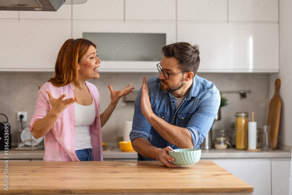 Intense disagreement between a couple in their kitchen. The man raises his hand in a defensive manner. Both display strong emotions, reflecting a challenging moment in their relationship Intense disagreement between a couple in their kitchen. The man raises his hand in a defensive manner. Both display strong emotions, reflecting a challenging moment in their relationship