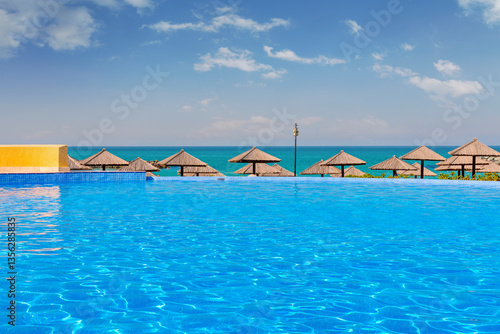 Obraz na płótnie Luxury hotel pool in Sharjah with crystal-clear water, surrounded by elegant sun loungers. Stunning sea views and a golden sandy beach create a relaxing atmosphere under the bright daytime sun. w salonie